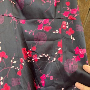 Op de afbeelding: Een donker marineblauwe jurk met een levendig bloemenpatroon met roze bloesems en witte accenten. Het ontwerp bevat gestileerde cirkelvormige en rechthoekige motieven, die een opvallend contrast cre&euml;ren met de donkere achtergrond. De jurk heeft een getailleerd lijfje en een uitlopende rok.