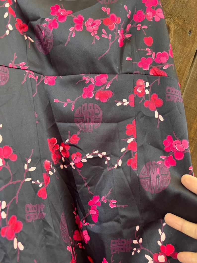 Op de afbeelding: Een donker marineblauwe jurk met een levendig bloemenpatroon met roze bloesems en witte accenten. Het ontwerp bevat gestileerde cirkelvormige en rechthoekige motieven, die een opvallend contrast cre&euml;ren met de donkere achtergrond. De jurk heeft een getailleerd lijfje en een uitlopende rok.