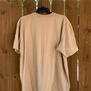 K&ouml;nnte beinhalten: Ein einfarbiges, hellbraunes T-Shirt mit kurzen &Auml;rmeln, das an einem schwarzen Kleiderb&uuml;gel h&auml;ngt. Das T-Shirt hat einen Rundhalsausschnitt und ist aus einem weichen Material. Im Hintergrund sind ein Holzzaun und gr&uuml;nes Laub zu sehen.