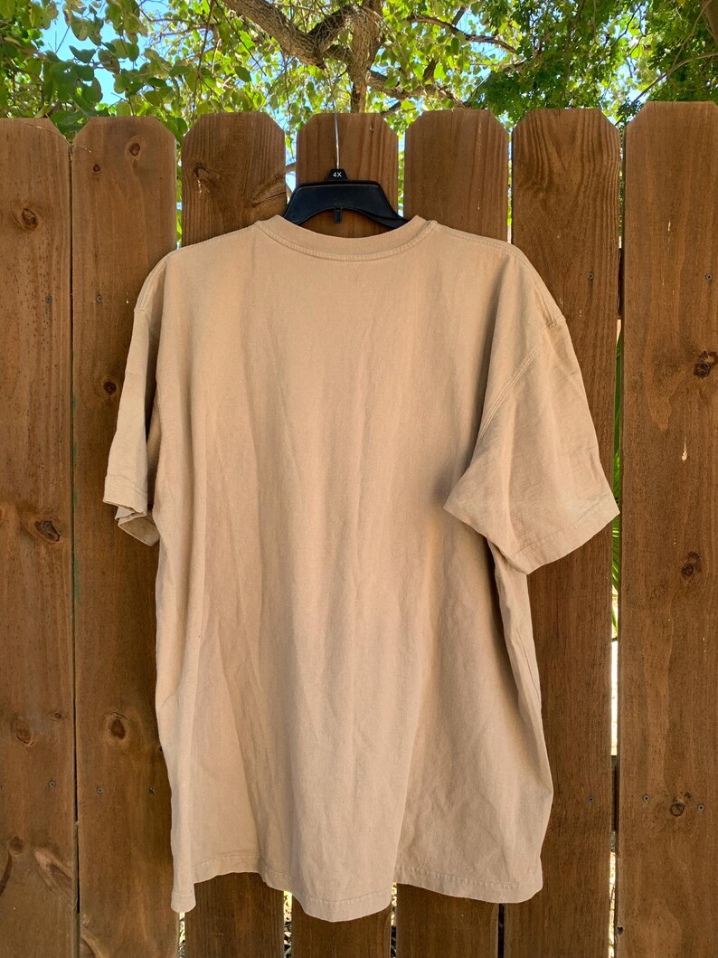 K&ouml;nnte beinhalten: Ein einfarbiges, hellbraunes T-Shirt mit kurzen &Auml;rmeln, das an einem schwarzen Kleiderb&uuml;gel h&auml;ngt. Das T-Shirt hat einen Rundhalsausschnitt und ist aus einem weichen Material. Im Hintergrund sind ein Holzzaun und gr&uuml;nes Laub zu sehen.
