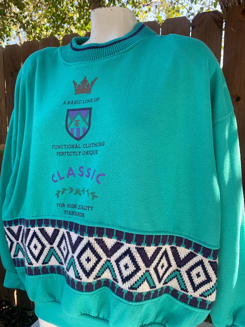 Puede incluir: Sudadera de color turquesa con un estampado geom&eacute;trico en blanco, turquesa y morado. El texto en la sudadera dice "A BASIC LINE OF FUNCTIONAL CLOTHING PERFECTLY UNIQUE CLASSIC FOR HIGH QUALITY STANDARDS". El cuello es turquesa con una raya azul marino.