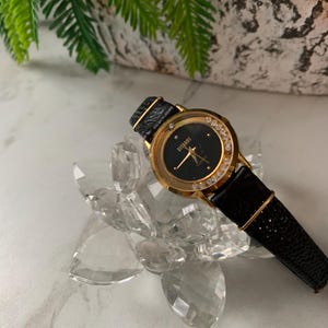 May include: A gold-tone wristwatch with a black face and a black leather band. The watch face has a diamond-like design around the edge and the word "CITIZEN" printed on it.