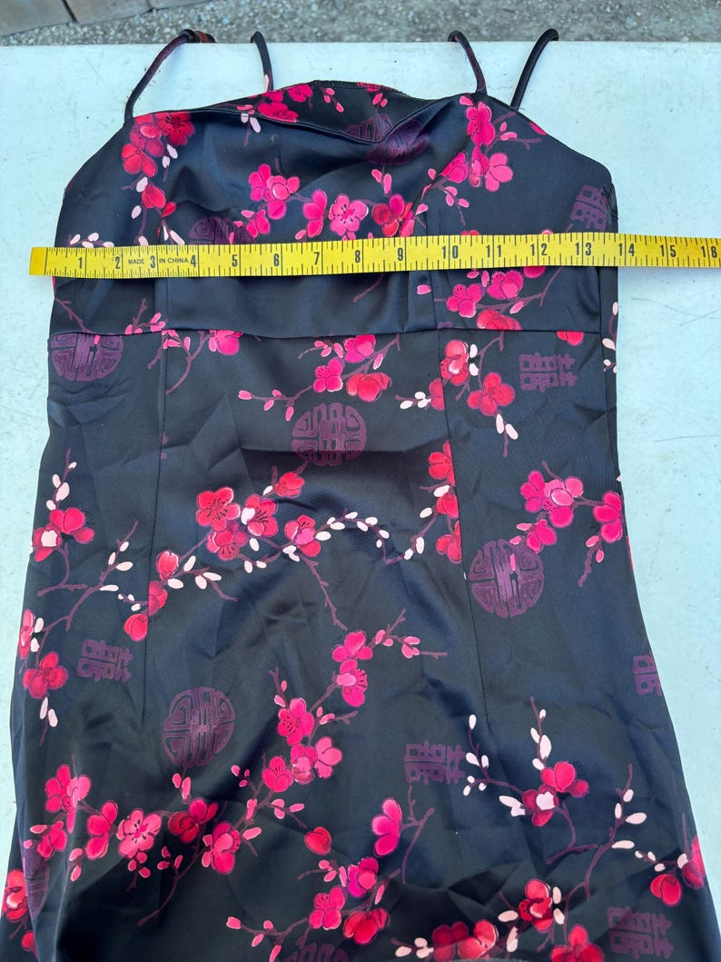 Op de afbeelding: Zwarte satijnen jurk met spaghettibandjes, versierd met een levendig bloemenpatroon in roze en rode tinten. De jurk heeft een getailleerd lijfje en een rechte rok. De jurk is ongeveer 35.5 cm breed.