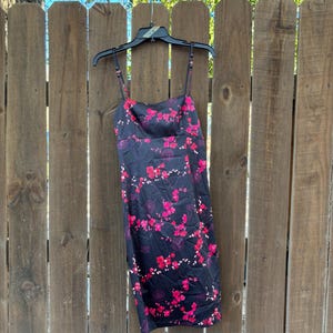 Op de afbeelding: Een zwarte slipdress met een levendig roze bloemenpatroon. De jurk heeft dunne spaghettibandjes en een vierkante halslijn. Het hangt aan een zwarte hanger tegen een houten hek.