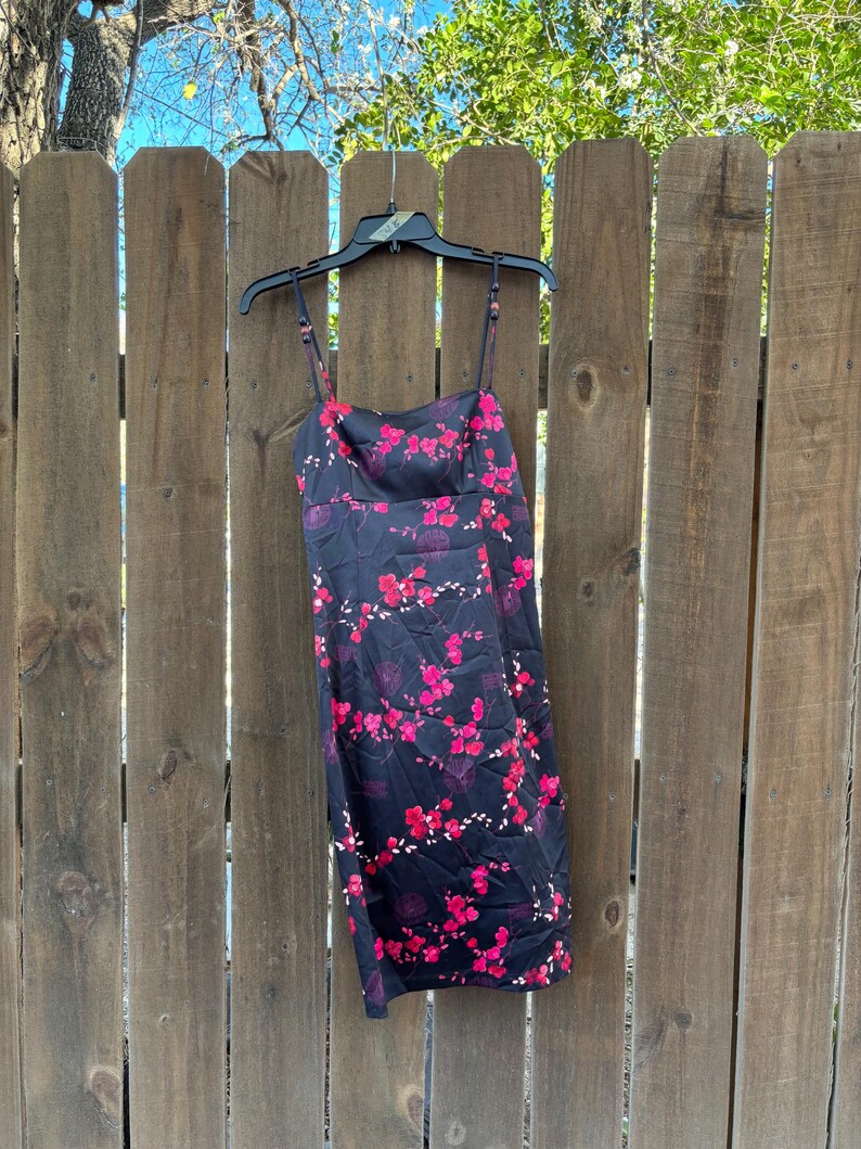 Op de afbeelding: Een zwarte slipdress met een levendig roze bloemenpatroon. De jurk heeft dunne spaghettibandjes en een vierkante halslijn. Het hangt aan een zwarte hanger tegen een houten hek.