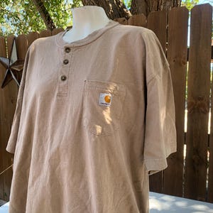 K&ouml;nnte beinhalten: Ein hellbraunes Kurzarm-Shirt mit Henley-Ausschnitt und Brusttasche. Das Shirt hat drei braune Kn&ouml;pfe und ein kleines Carhartt-Logo auf der Tasche. Das Shirt wird auf einer Schaufensterpuppe pr&auml;sentiert.