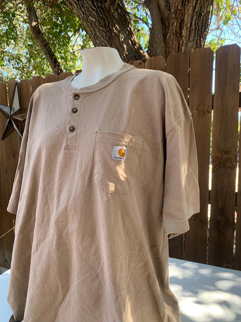 K&ouml;nnte beinhalten: Ein hellbraunes Kurzarm-Shirt mit Henley-Ausschnitt und Brusttasche. Das Shirt hat drei braune Kn&ouml;pfe und ein kleines Carhartt-Logo auf der Tasche. Das Shirt wird auf einer Schaufensterpuppe pr&auml;sentiert.