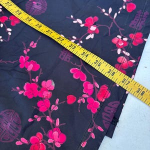 Op de afbeelding: Zwarte stof met een levendig bloemmotief van roze bloesems en witte bladeren. Het ontwerp bevat traditionele Chinese karakters en cirkelvormige motieven. Een gele meetlint ligt over de stof en toont de afmetingen in centimeters.
