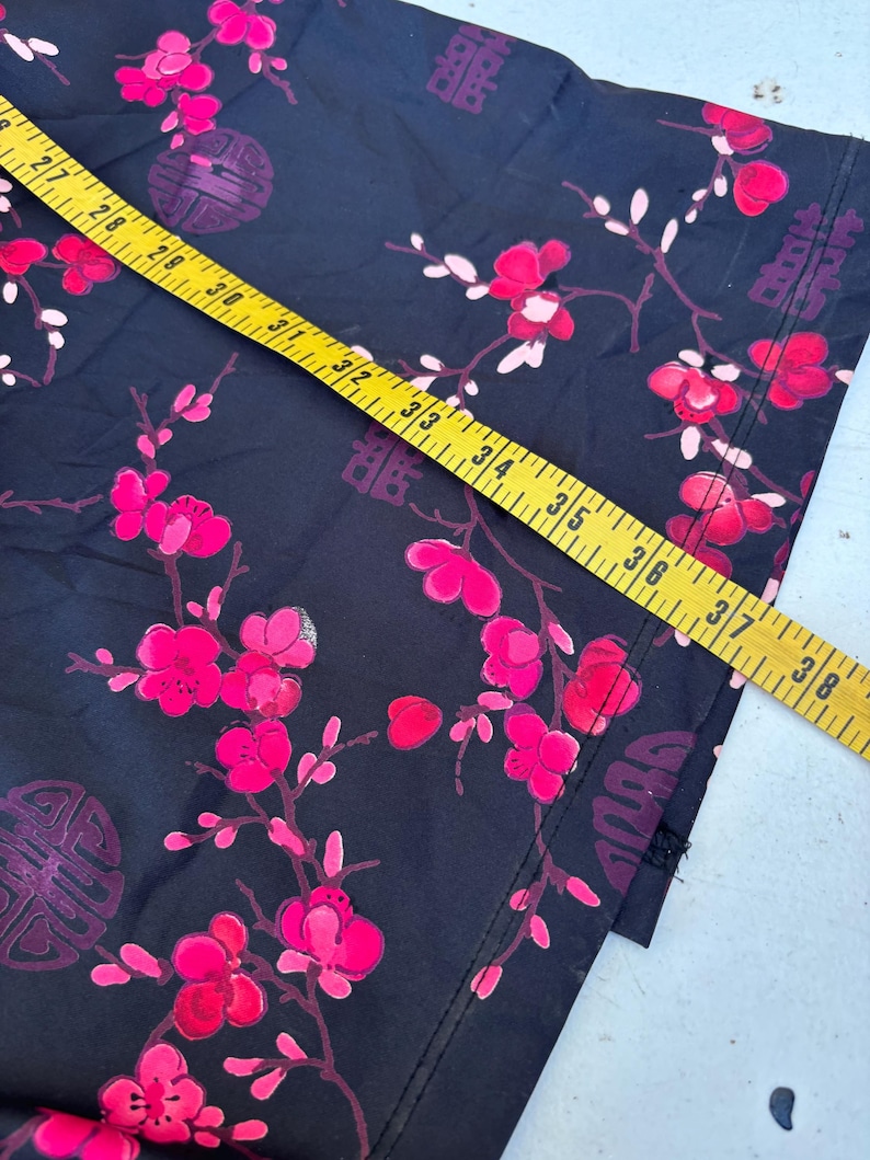 Op de afbeelding: Zwarte stof met een levendig bloemmotief van roze bloesems en witte bladeren. Het ontwerp bevat traditionele Chinese karakters en cirkelvormige motieven. Een gele meetlint ligt over de stof en toont de afmetingen in centimeters.