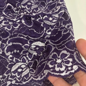 May include: Close-up of a piece of purple lace fabric with a floral pattern. The lace has a delicate, intricate design with white accents, creating a contrast against the deep purple background. The edge of the fabric is scalloped.