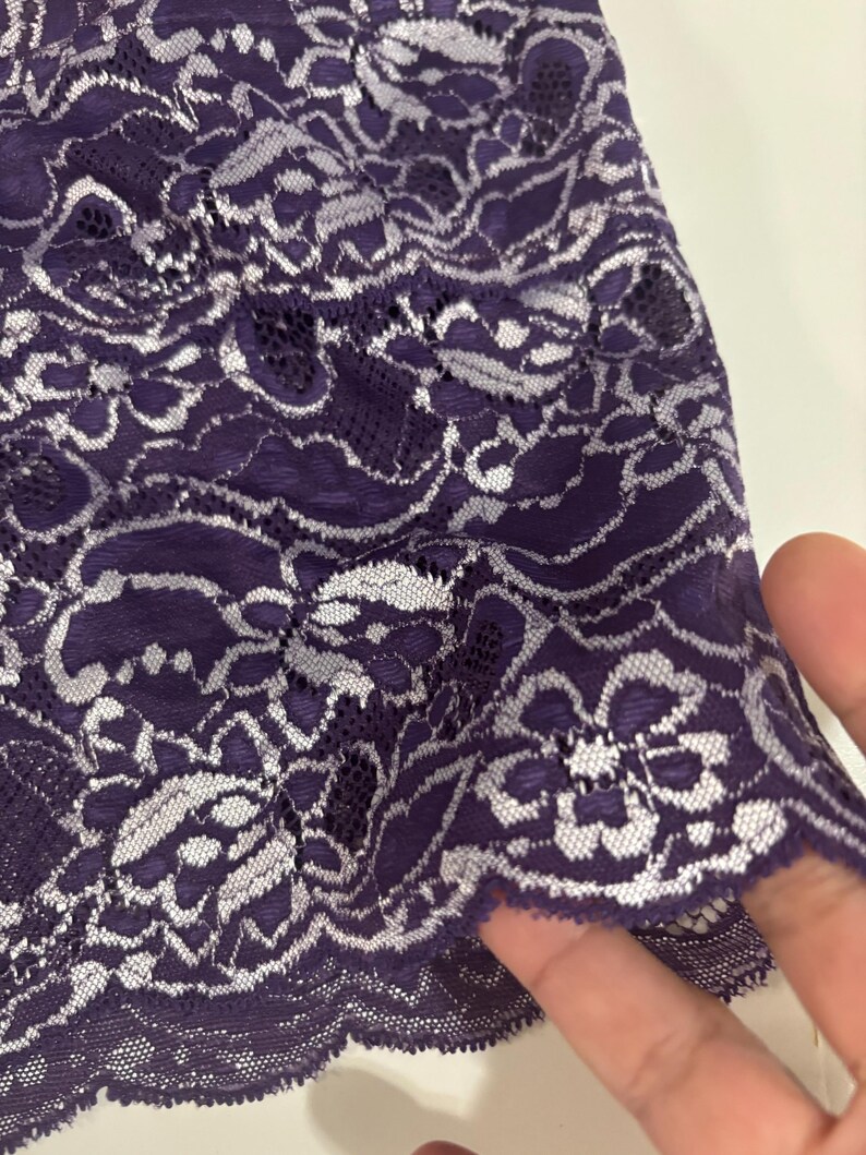 May include: Close-up of a piece of purple lace fabric with a floral pattern. The lace has a delicate, intricate design with white accents, creating a contrast against the deep purple background. The edge of the fabric is scalloped.