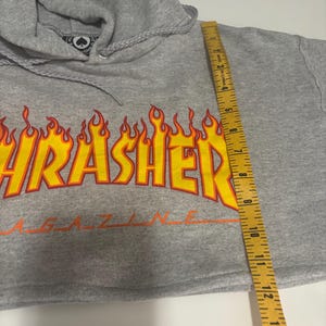 May include: A gray cropped hoodie with a drawstring hood. The front features a large, flame-style graphic with the word "Thrasher" in yellow and orange. The hoodie is measured with a 14-inch ruler.