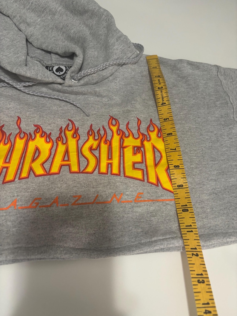 May include: A gray cropped hoodie with a drawstring hood. The front features a large, flame-style graphic with the word "Thrasher" in yellow and orange. The hoodie is measured with a 14-inch ruler.