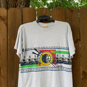 May include: A gray t-shirt with a colorful graphic print of rowers on a boat. The graphic includes a globe and the text "St. John's Bay."