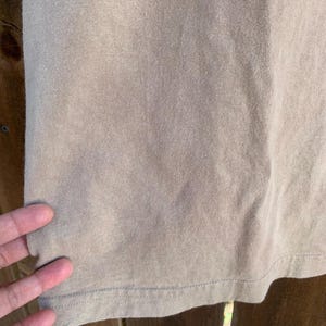 K&ouml;nnte beinhalten: Nahaufnahme eines tan-farbenen T-Shirts. Der Stoff hat eine gerippte Textur und einen sichtbaren Saum mit N&auml;hten. Das Shirt scheint aus einem weichen, bequemen Material zu bestehen. Das Bild ist gut beleuchtet und hebt die Details des Stoffes hervor.