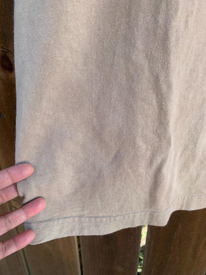 K&ouml;nnte beinhalten: Nahaufnahme eines tan-farbenen T-Shirts. Der Stoff hat eine gerippte Textur und einen sichtbaren Saum mit N&auml;hten. Das Shirt scheint aus einem weichen, bequemen Material zu bestehen. Das Bild ist gut beleuchtet und hebt die Details des Stoffes hervor.