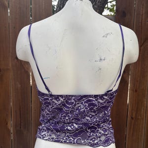 May include: A purple lace camisole with thin, adjustable straps. The camisole is displayed on a white mannequin against a wooden fence background. The lace pattern is intricate and detailed.