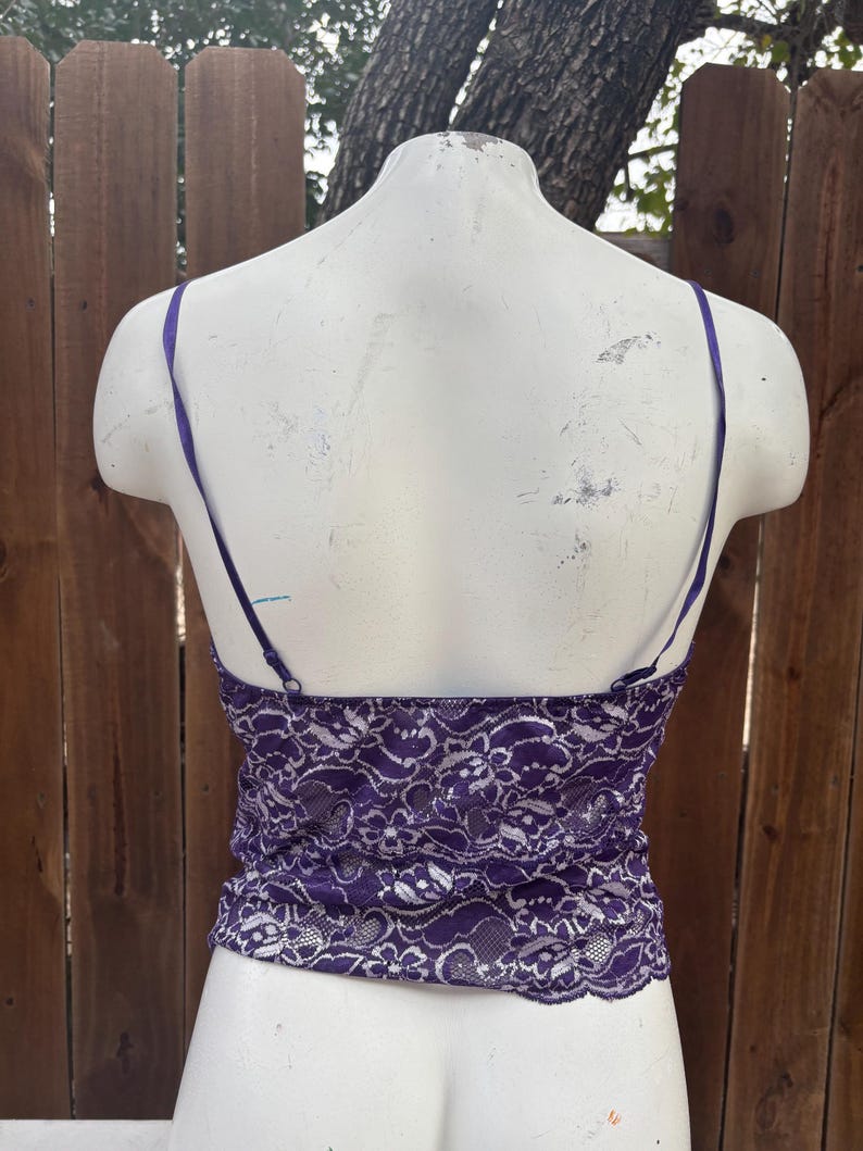 May include: A purple lace camisole with thin, adjustable straps. The camisole is displayed on a white mannequin against a wooden fence background. The lace pattern is intricate and detailed.