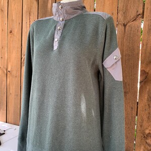 May include: A green long-sleeved sweater with a gray collar and a button closure. The sweater has a patch pocket on the left sleeve.