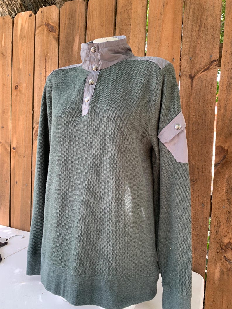 May include: A green long-sleeved sweater with a gray collar and a button closure. The sweater has a patch pocket on the left sleeve.