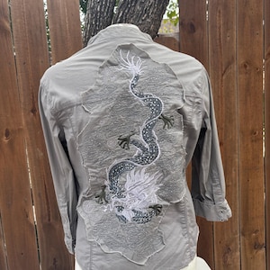 K&ouml;nnte beinhalten: Graues Hemd mit Kn&ouml;pfen und einem detaillierten, gestickten Drachendesign auf dem R&uuml;cken. Der Drache ist haupts&auml;chlich grau und wei&szlig;, mit aufwendigen Details. Das Hemd hat lange &Auml;rmel und einen klassischen Kragen.