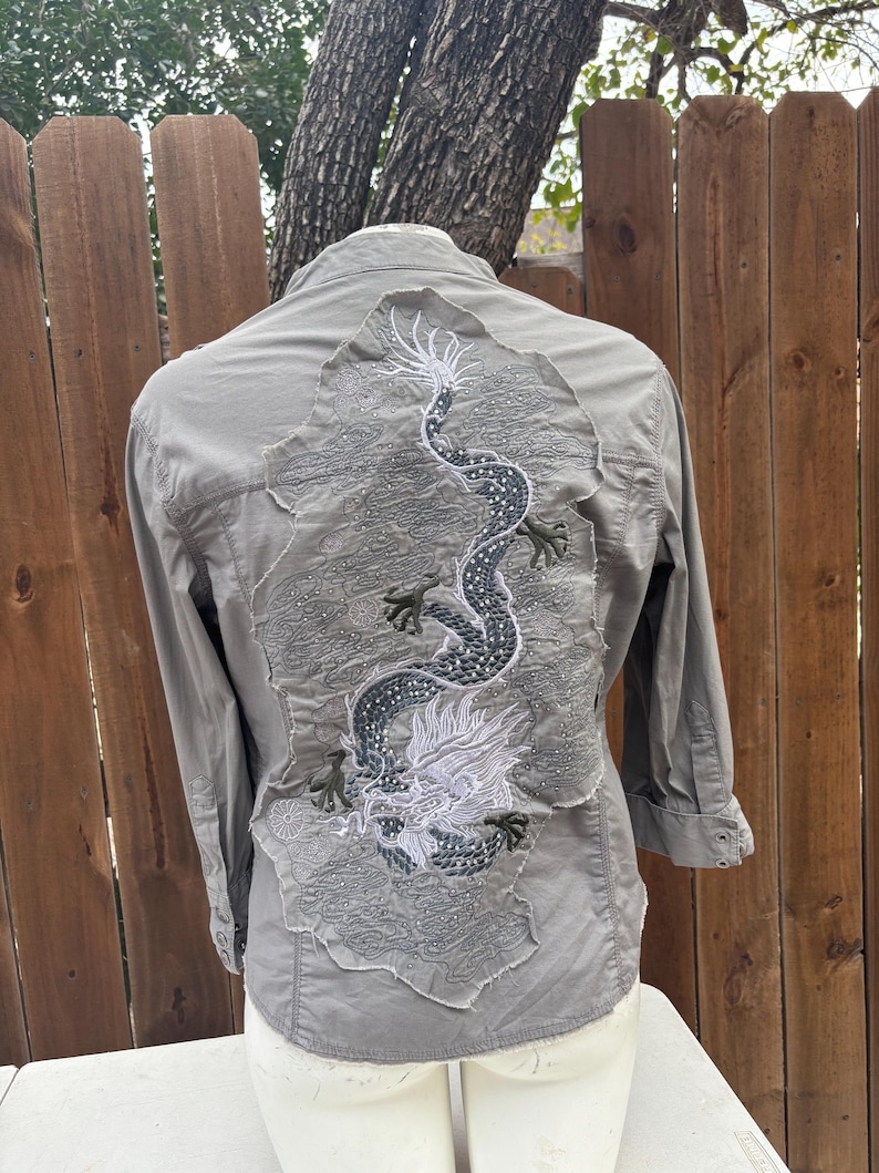 K&ouml;nnte beinhalten: Graues Hemd mit Kn&ouml;pfen und einem detaillierten, gestickten Drachendesign auf dem R&uuml;cken. Der Drache ist haupts&auml;chlich grau und wei&szlig;, mit aufwendigen Details. Das Hemd hat lange &Auml;rmel und einen klassischen Kragen.