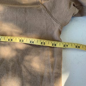 K&ouml;nnte beinhalten: Ein beigefarbenes Sweatshirt liegt flach, mit einem gelben Ma&szlig;band dar&uuml;ber. Das Band zeigt Messungen von 56 bis 71 cm. Der Stoff scheint ein weiches, gestricktes Material zu sein.