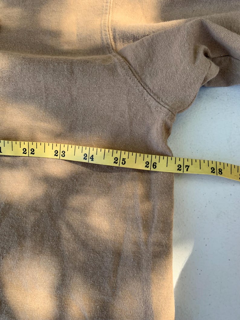K&ouml;nnte beinhalten: Ein beigefarbenes Sweatshirt liegt flach, mit einem gelben Ma&szlig;band dar&uuml;ber. Das Band zeigt Messungen von 56 bis 71 cm. Der Stoff scheint ein weiches, gestricktes Material zu sein.
