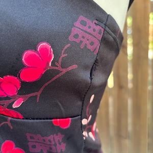 Op de afbeelding: Close-up van zwarte stof met een bloemenpatroon in roze en rood. Het ontwerp omvat gestileerde bloemen en Chinese karakters. De stof lijkt deel uit te maken van een kledingstuk, met zichtbare naden en bandjes.