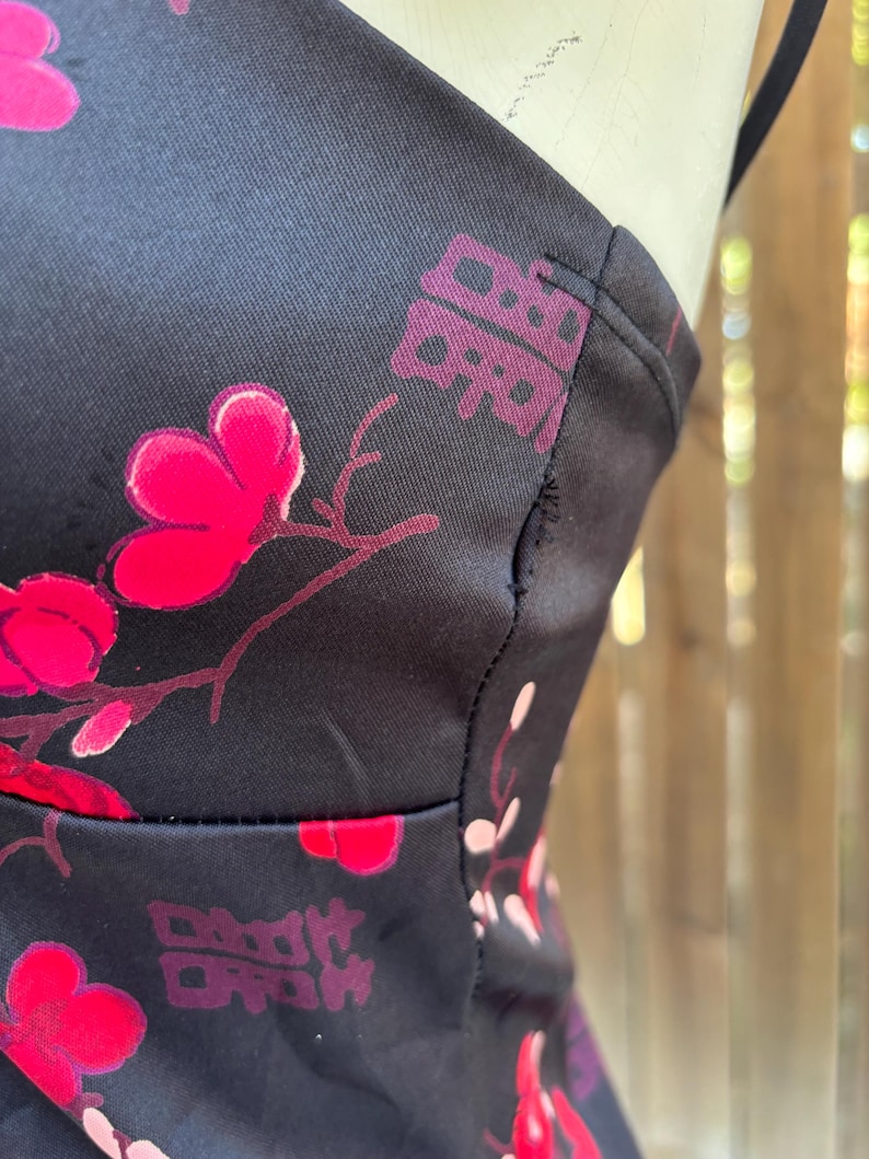 Op de afbeelding: Close-up van zwarte stof met een bloemenpatroon in roze en rood. Het ontwerp omvat gestileerde bloemen en Chinese karakters. De stof lijkt deel uit te maken van een kledingstuk, met zichtbare naden en bandjes.