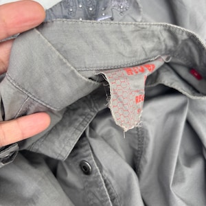 K&ouml;nnte beinhalten: Nahaufnahme eines grauen Button-Down-Hemds. Der Kragen hat ein gesticktes Muster und kleine Verzierungen. Das Hemd hat schwarze Kn&ouml;pfe und ein rotes Etikett mit Text.