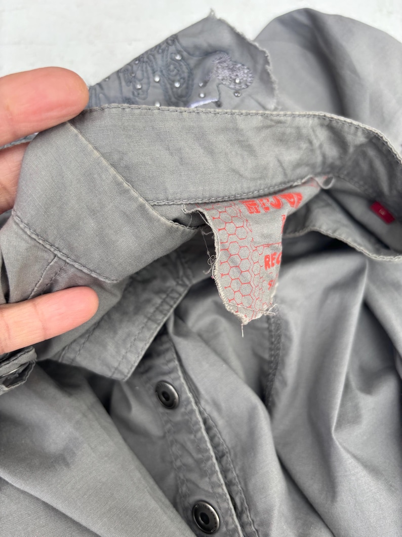 K&ouml;nnte beinhalten: Nahaufnahme eines grauen Button-Down-Hemds. Der Kragen hat ein gesticktes Muster und kleine Verzierungen. Das Hemd hat schwarze Kn&ouml;pfe und ein rotes Etikett mit Text.