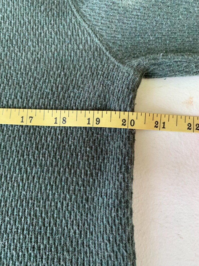 May include: Close-up of a dark green fabric with a textured pattern. A measuring tape is placed along the edge of the fabric, showing measurements from 17 to 22 inches.