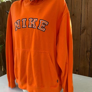 Peut inclure: Un sweat &agrave; capuche orange Nike avec une poche avant et une capuche. Le mot "NIKE" est imprim&eacute; sur la poitrine en lettres blanches avec un contour noir. L'int&eacute;rieur de la capuche est bleu marine.