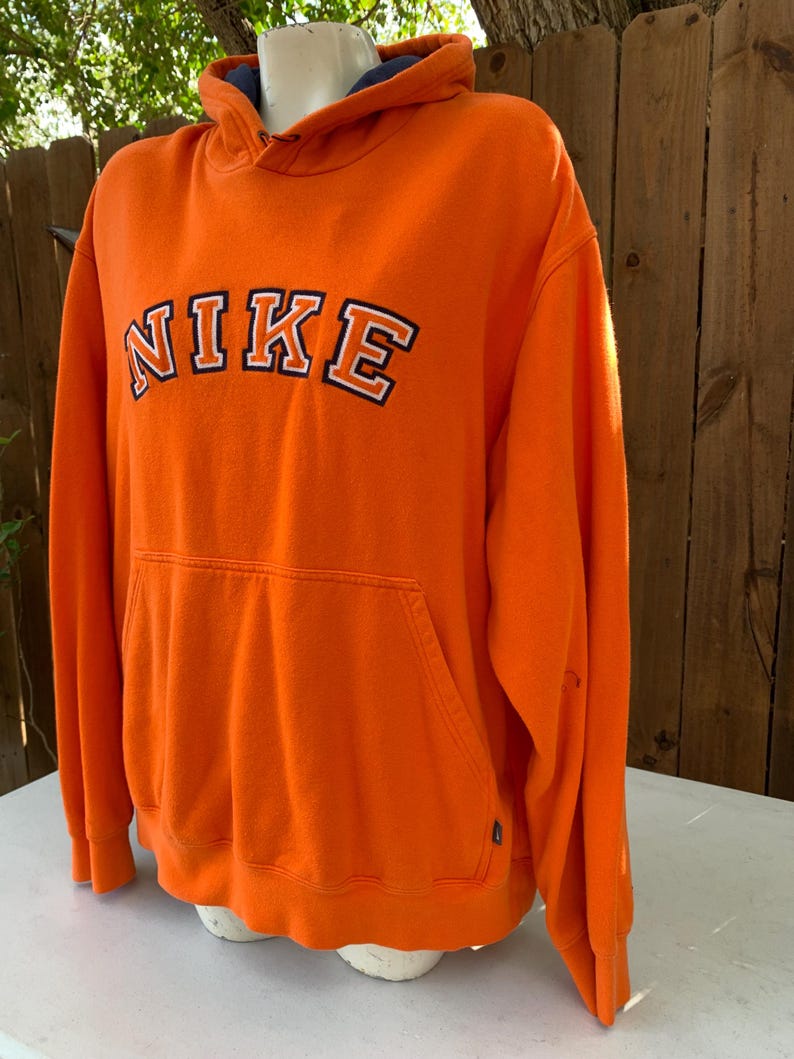 Peut inclure: Un sweat &agrave; capuche orange Nike avec une poche avant et une capuche. Le mot "NIKE" est imprim&eacute; sur la poitrine en lettres blanches avec un contour noir. L'int&eacute;rieur de la capuche est bleu marine.