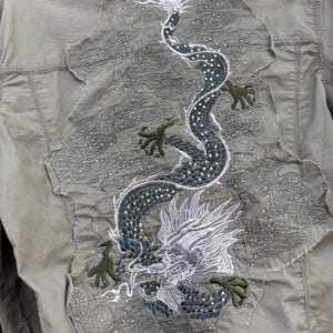 K&ouml;nnte beinhalten: Hell olivgr&uuml;ne Jacke mit gro&szlig;em, gesticktem Drachendesign auf dem R&uuml;cken. Der Drache ist haupts&auml;chlich dunkelgrau und wei&szlig;, mit gr&uuml;nen Akzenten. Das Design befindet sich auf einem Patch mit ausgefranstem Rand.
