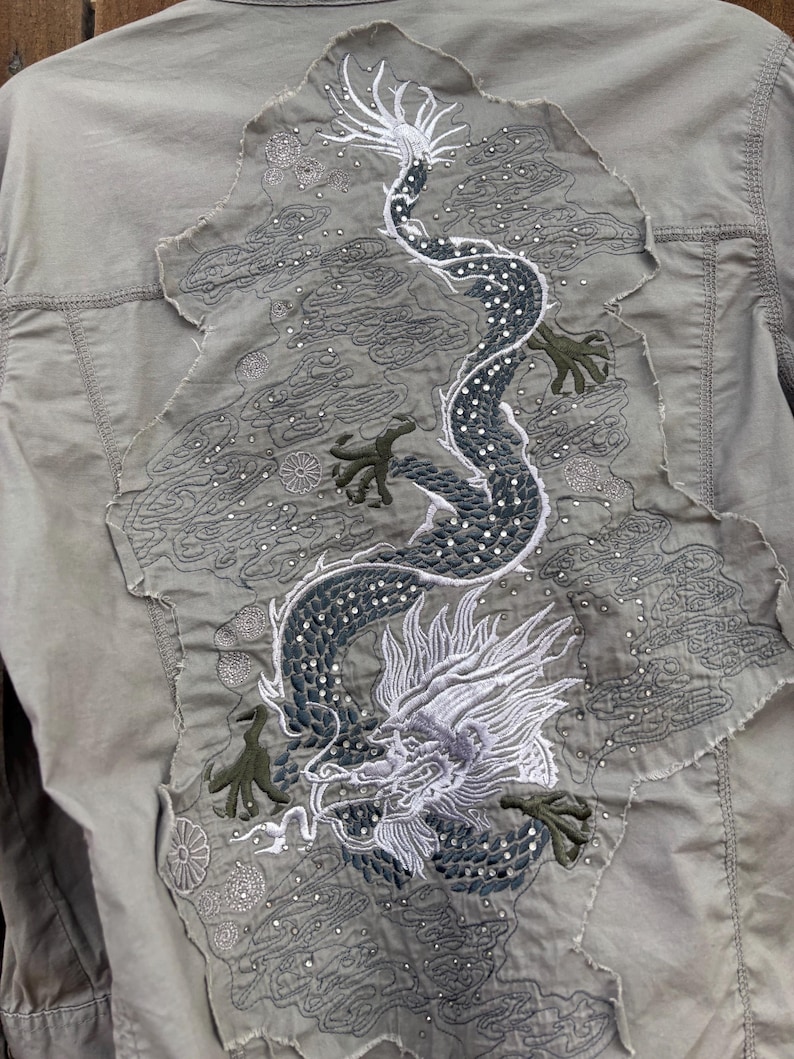 K&ouml;nnte beinhalten: Hell olivgr&uuml;ne Jacke mit gro&szlig;em, gesticktem Drachendesign auf dem R&uuml;cken. Der Drache ist haupts&auml;chlich dunkelgrau und wei&szlig;, mit gr&uuml;nen Akzenten. Das Design befindet sich auf einem Patch mit ausgefranstem Rand.