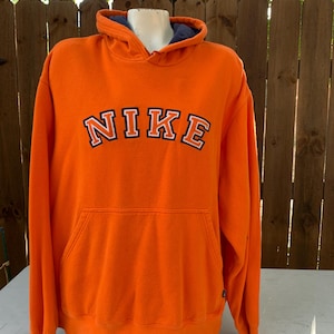 Peut inclure: Un sweat &agrave; capuche orange vif avec le mot "NIKE" en lettres blanches et noires sur la poitrine. Le sweat &agrave; capuche a une poche avant et une doublure bleue &agrave; l'int&eacute;rieur de la capuche. Le v&ecirc;tement est pr&eacute;sent&eacute; sur un mannequin.