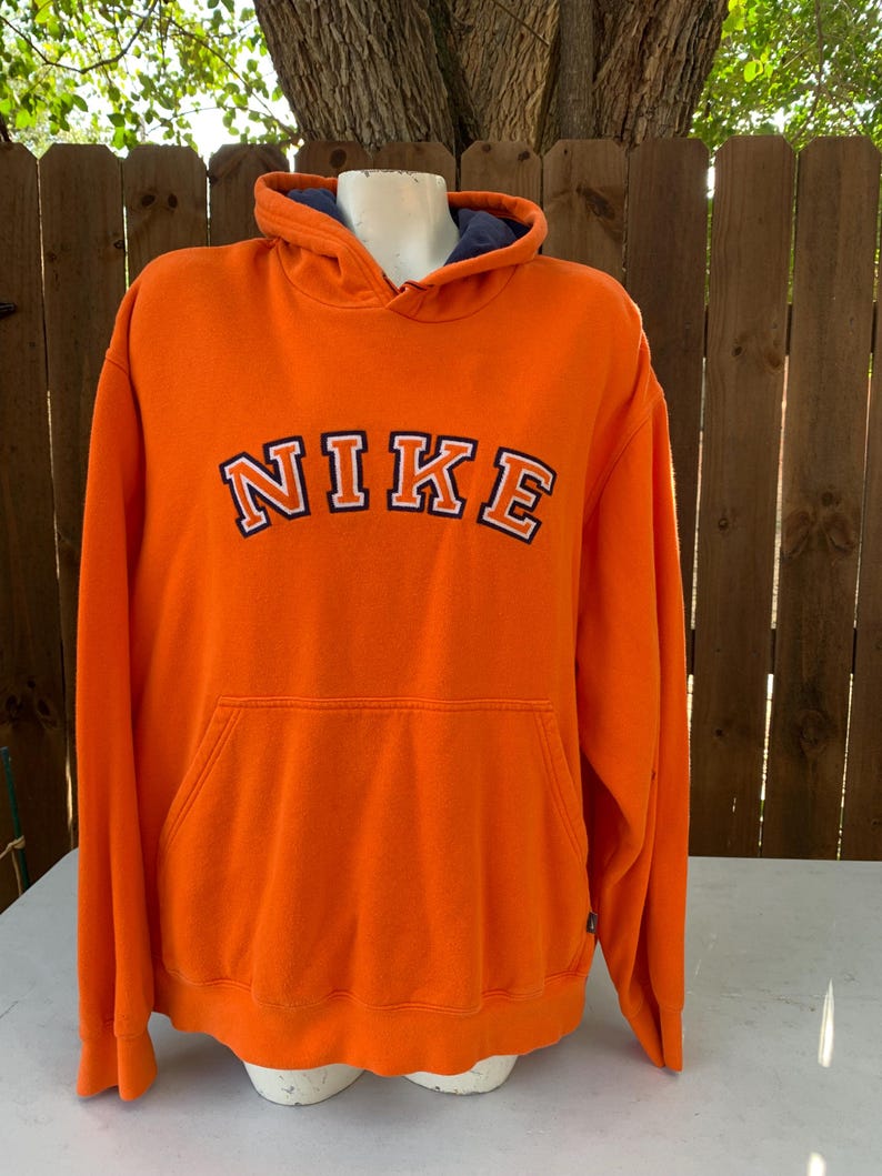 Peut inclure: Un sweat &agrave; capuche orange vif avec le mot "NIKE" en lettres blanches et noires sur la poitrine. Le sweat &agrave; capuche a une poche avant et une doublure bleue &agrave; l'int&eacute;rieur de la capuche. Le v&ecirc;tement est pr&eacute;sent&eacute; sur un mannequin.