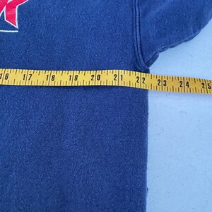 May include: Navy blue sweatshirt with a red and white graphic on the upper left. A yellow measuring tape is laid across the garment, showing a measurement of approximately 22 inches.