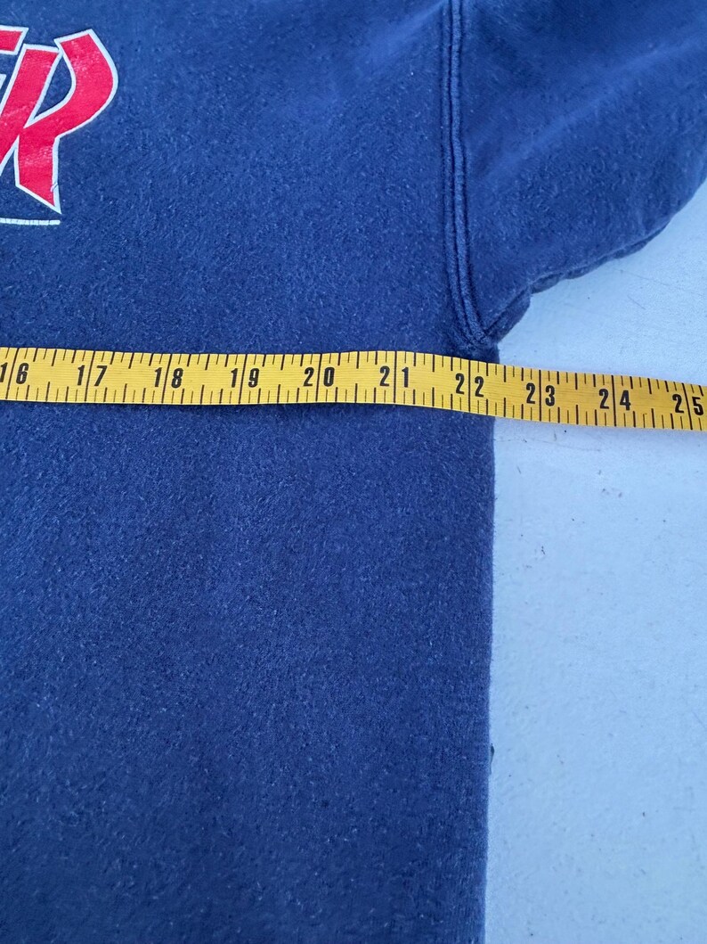 May include: Navy blue sweatshirt with a red and white graphic on the upper left. A yellow measuring tape is laid across the garment, showing a measurement of approximately 22 inches.