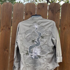 K&ouml;nnte beinhalten: Hellolivgr&uuml;nes Hemd mit detaillierter Drachenstickerei auf dem R&uuml;cken. Der Drache ist haupts&auml;chlich grau und wei&szlig;, mit gr&uuml;nen Akzenten. Das Hemd hat lange &Auml;rmel und einen klassischen Kragen, pr&auml;sentiert auf einem Holzzaun.