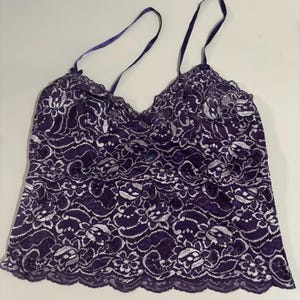 May include: A purple lace camisole with thin, dark purple straps. The camisole features a scalloped edge and a delicate floral lace pattern. The fabric is a deep purple with white accents, creating a contrasting design.