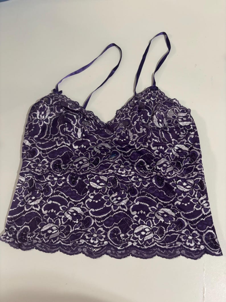 May include: A purple lace camisole with thin, dark purple straps. The camisole features a scalloped edge and a delicate floral lace pattern. The fabric is a deep purple with white accents, creating a contrasting design.