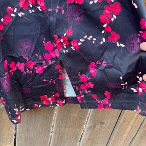 Op de afbeelding: Donker marineblauwe stof met een bloemenpatroon van rode en roze bloesems en witte accenten. Het ontwerp bevat cirkelvormige paarse symbolen. De stof is over een houten oppervlak gedrapeerd.