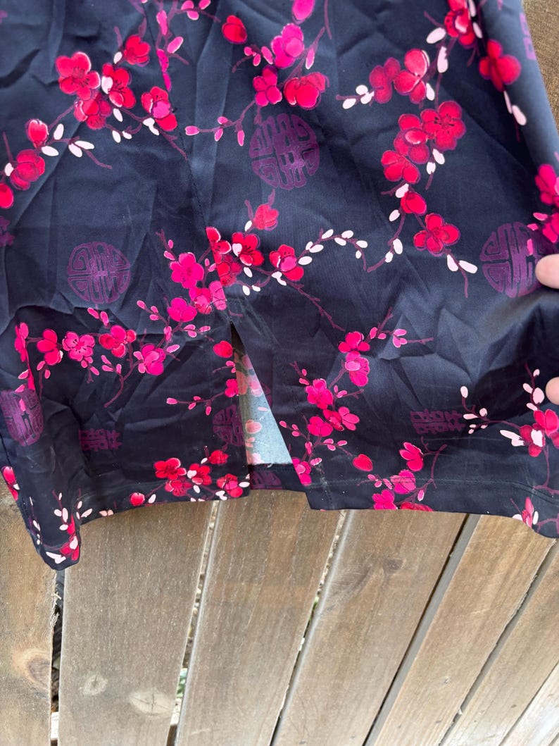 Op de afbeelding: Donker marineblauwe stof met een bloemenpatroon van rode en roze bloesems en witte accenten. Het ontwerp bevat cirkelvormige paarse symbolen. De stof is over een houten oppervlak gedrapeerd.