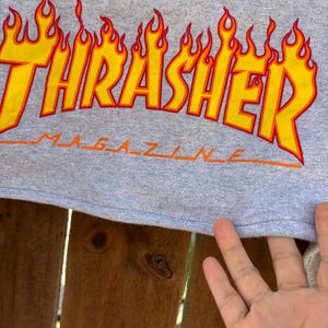 May include: Gray t-shirt featuring the Thrasher Magazine logo in yellow and orange flames. The word "Magazine" is printed in orange below the main logo. The shirt is made of a soft, comfortable material.