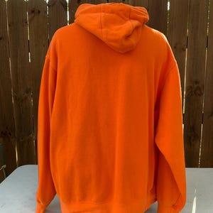 Peut inclure: Sweat-shirt &agrave; capuche orange vif. La vue arri&egrave;re montre la capuche et toute la longueur du sweat-shirt. Le sweat-shirt est fait d'une mati&egrave;re douce et poss&egrave;de une poche kangourou. L'arri&egrave;re-plan comprend une cl&ocirc;ture en bois et un arbre.