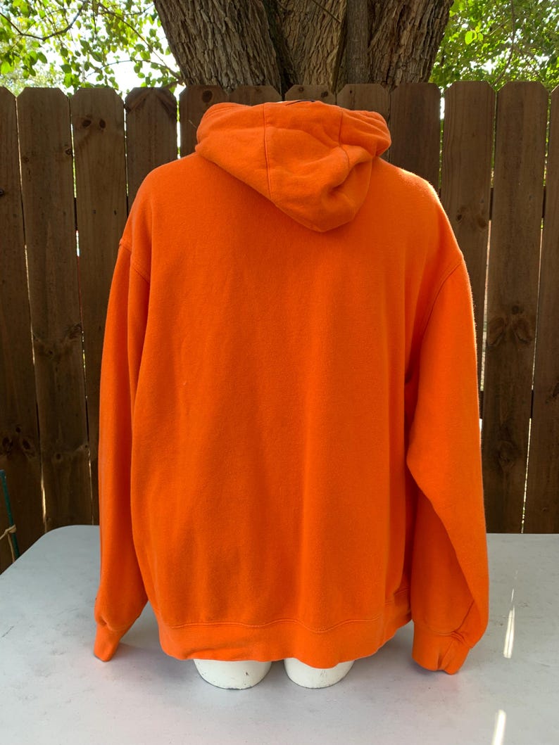 Peut inclure: Sweat-shirt &agrave; capuche orange vif. La vue arri&egrave;re montre la capuche et toute la longueur du sweat-shirt. Le sweat-shirt est fait d'une mati&egrave;re douce et poss&egrave;de une poche kangourou. L'arri&egrave;re-plan comprend une cl&ocirc;ture en bois et un arbre.