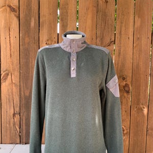 May include: A green long-sleeved sweater with a mock neck and button closure. The sweater has a gray patch on the shoulder and a gray patch on the left sleeve.